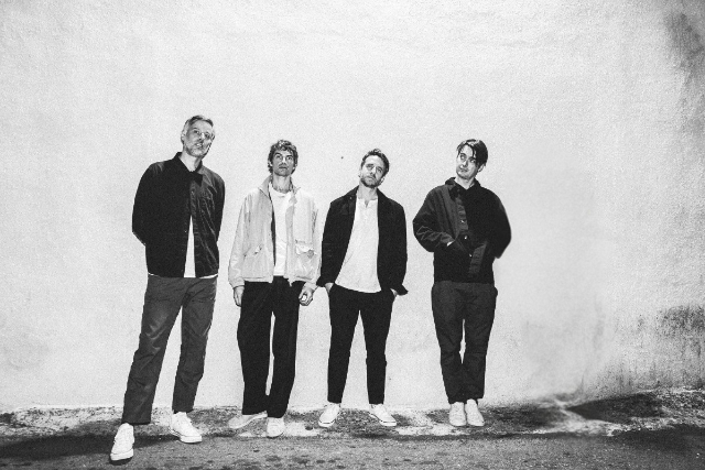 Four men stand against a white wall, wearing casual jackets and pants. Black and white photo.