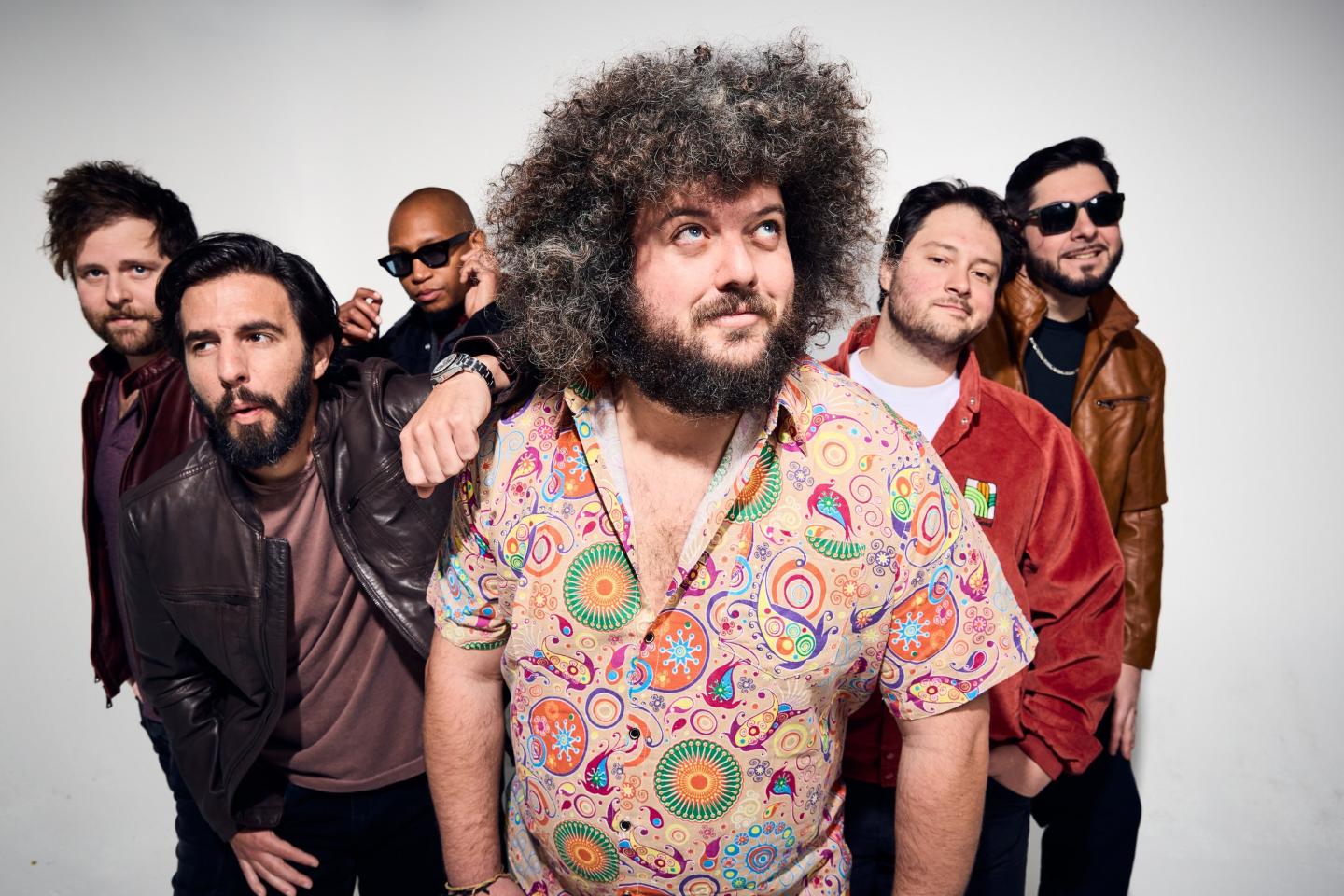 Group of six men posing, one with curly hair and colorful shirt, others in jackets.