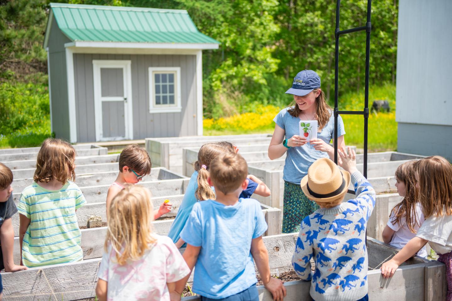 Children gather around a woman in a garden setting on a sunny day.