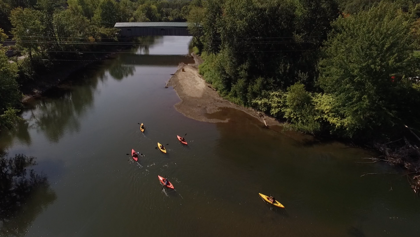 Kayakers paddling down a tree-lined river under a covered bridge.