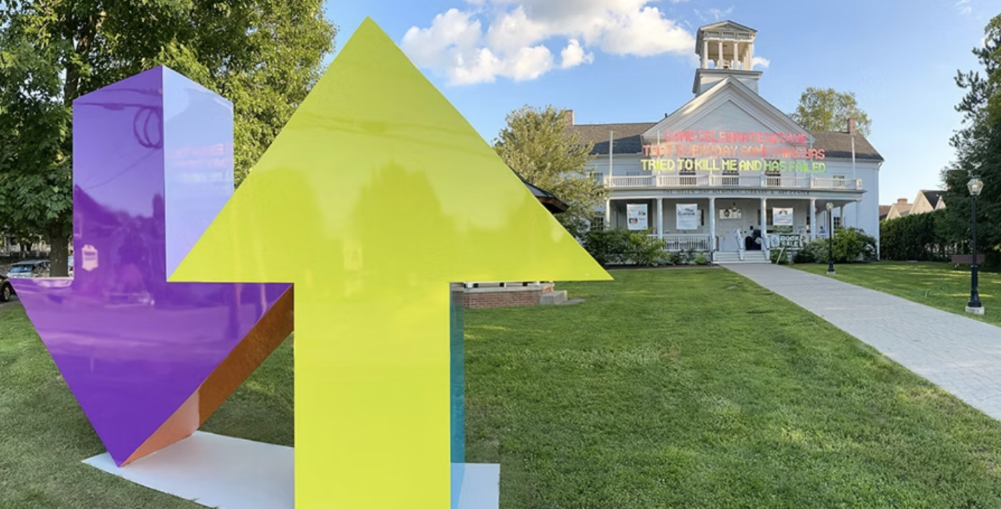 Bright yellow upward arrow and purple downward arrow sculpture on grass, with building in background.