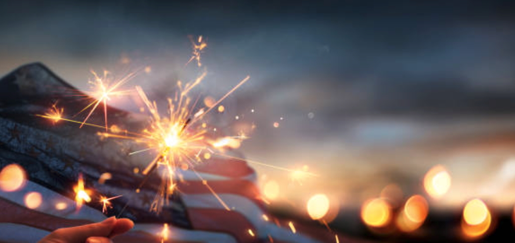Sparkler in hand, American flag backdrop, blurred lights in evening sky.