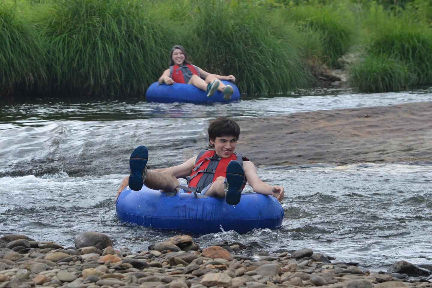 Two people tubing on a river, wearing life vests, surrounded by greenery.
