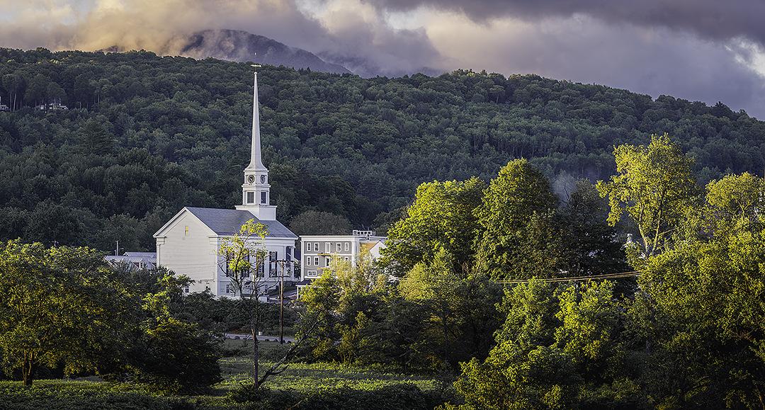 Church with tall steeple amidst lush greenery and cloudy mountain in the background.