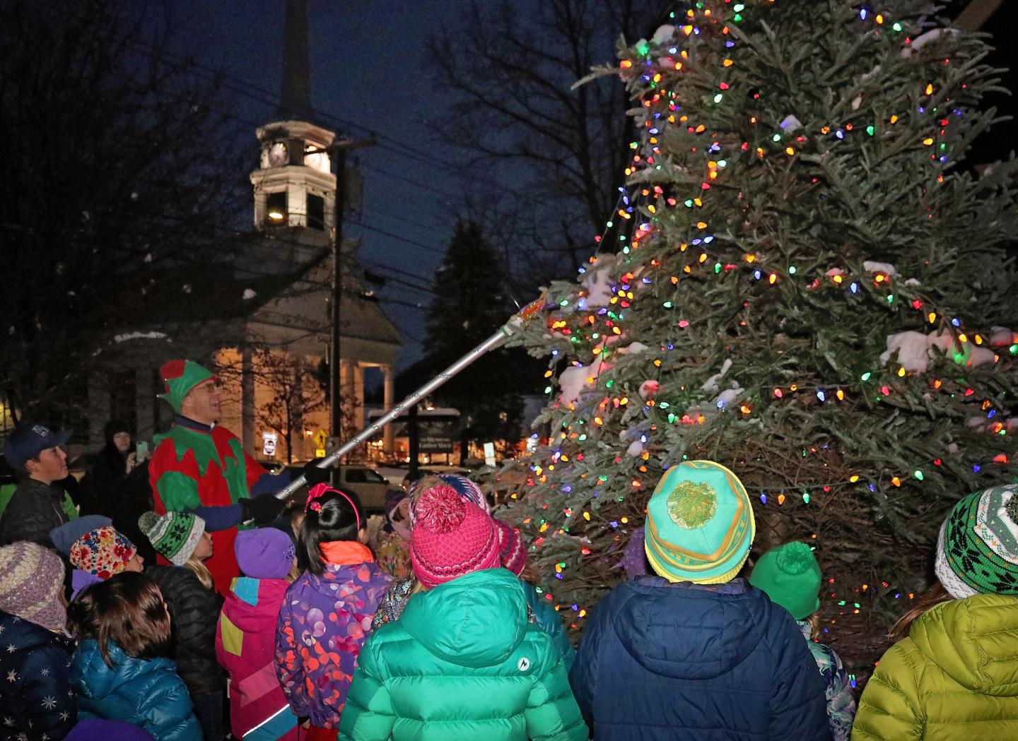 Children gather around a lit Christmas tree at night.