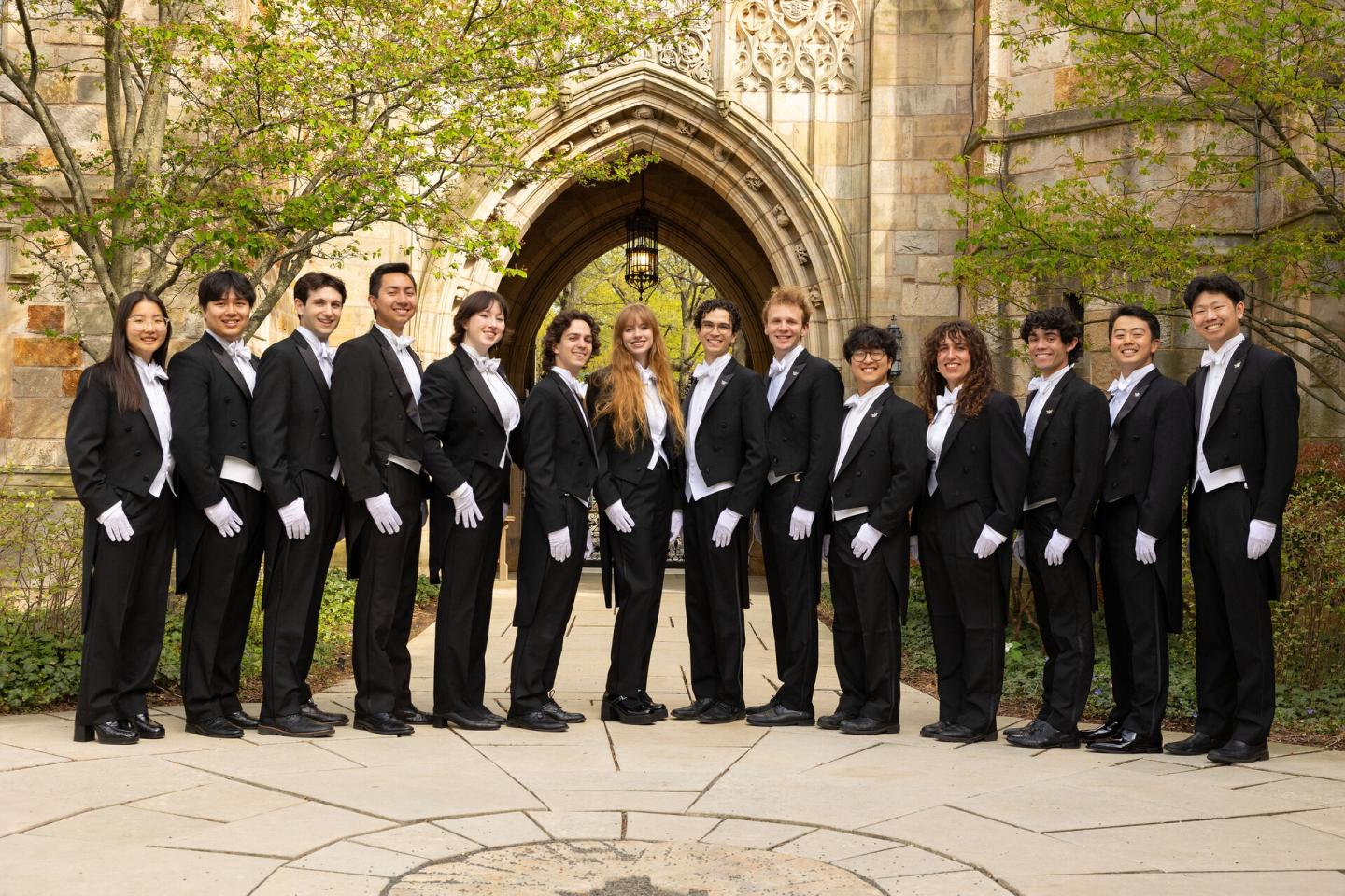 A group in black tuxedos stands in front of an ornate stone archway.