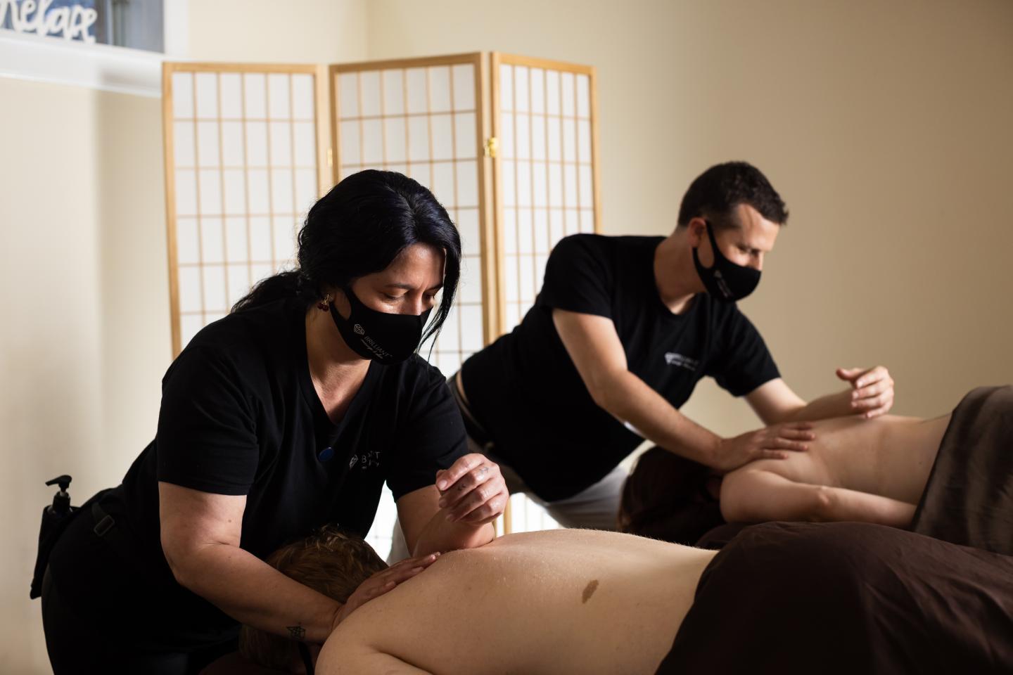 Two masked massage therapists working on clients in a serene room.