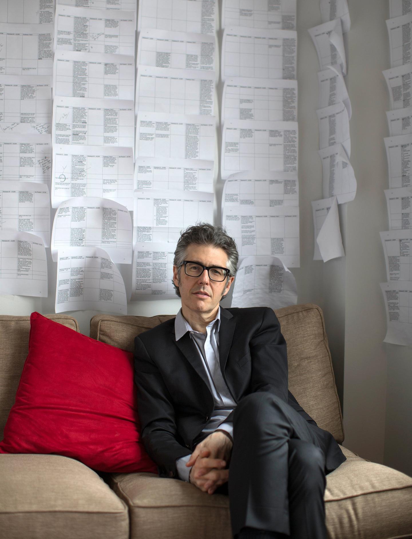 Man in a suit sitting on a sofa against a wall covered in paper sheets.