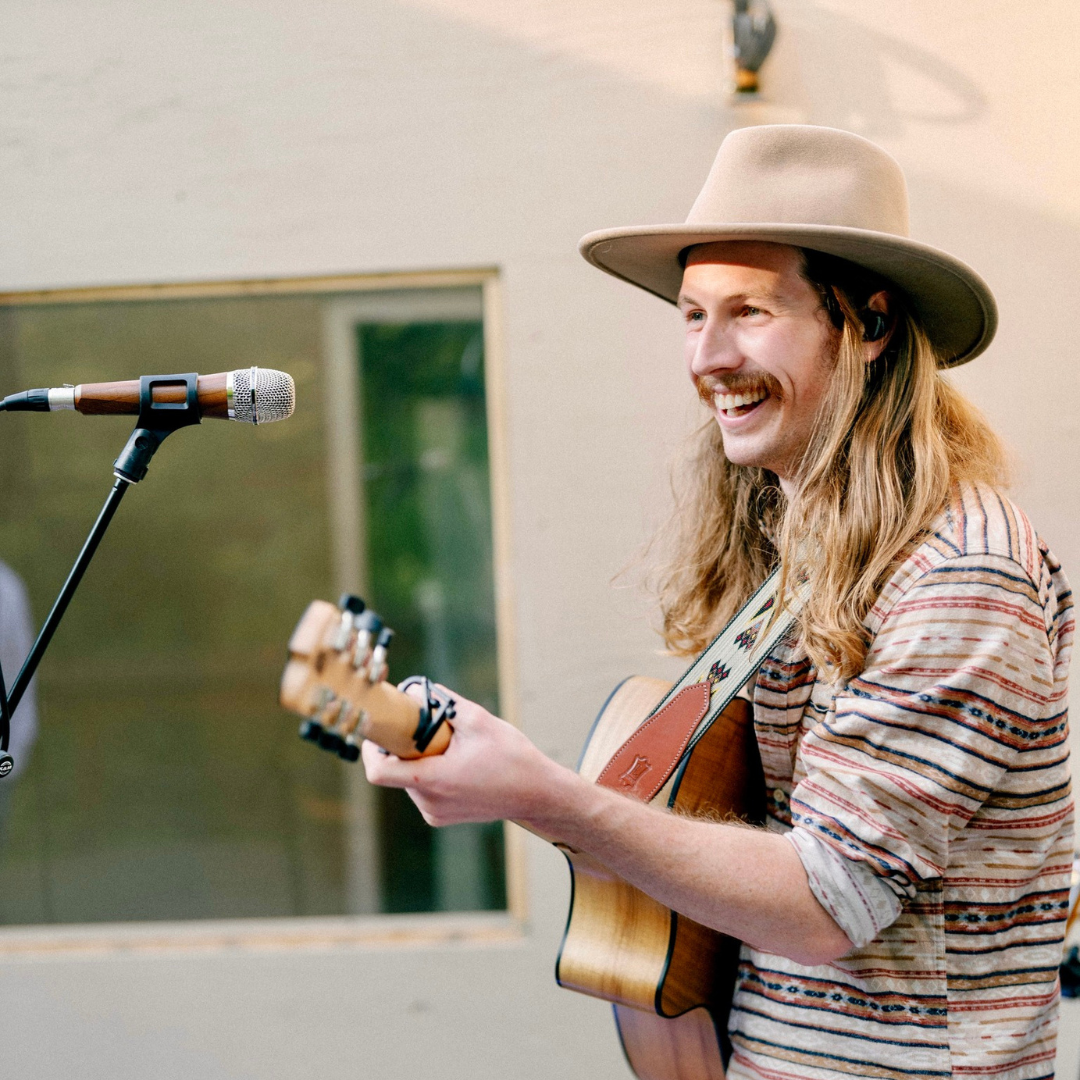 Musician with long hair plays guitar and sings joyfully into a microphone.