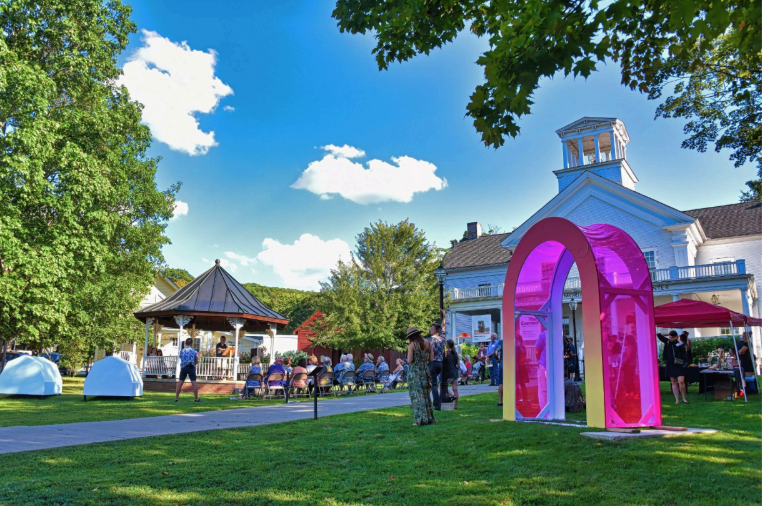 Outdoor art festival with colorful arch, gazebo, and white building under a blue sky.