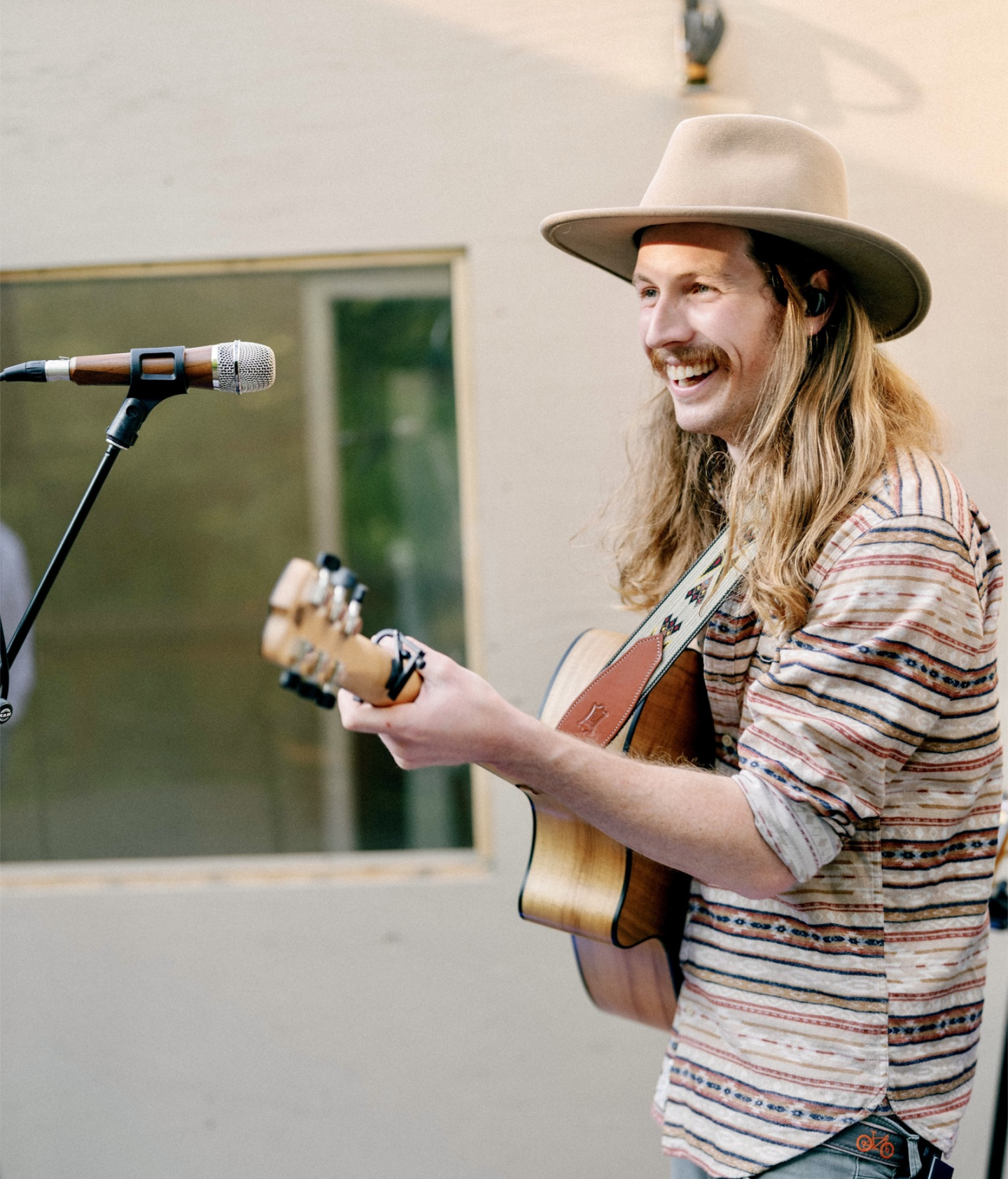 Musician with a guitar smiling near a microphone, wearing a hat and patterned shirt.