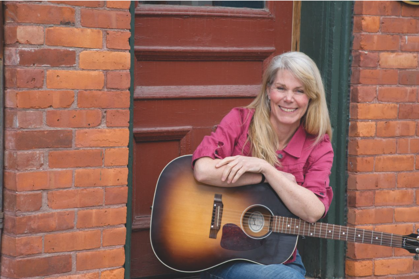 Smiling woman with a guitar, sitting by a red brick wall.