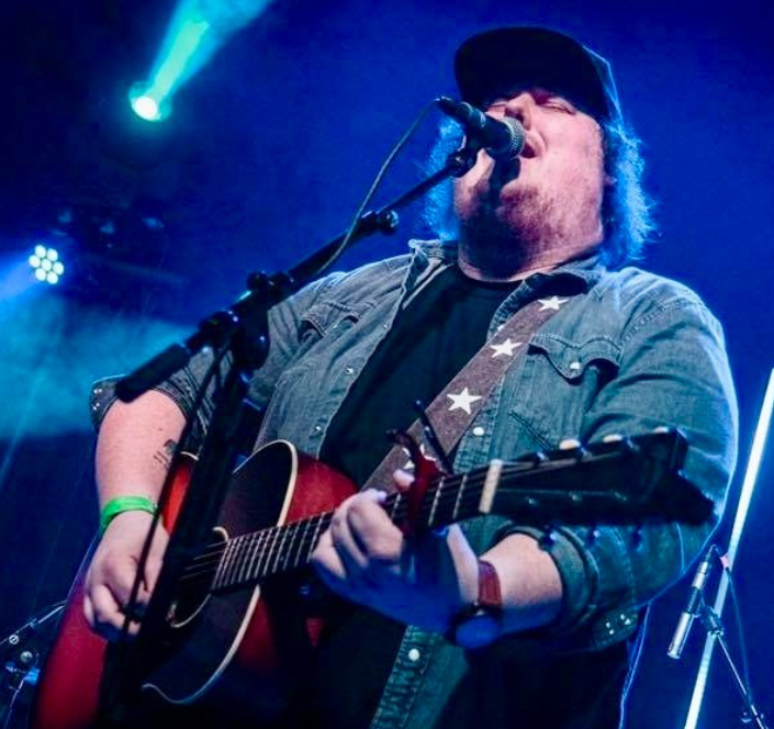 Musician in cap singing and playing guitar on stage with colorful lights.