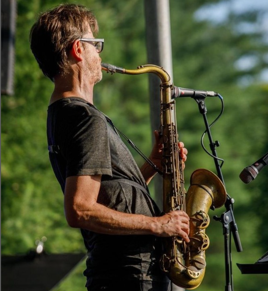 Musician playing saxophone outdoors, wearing sunglasses.