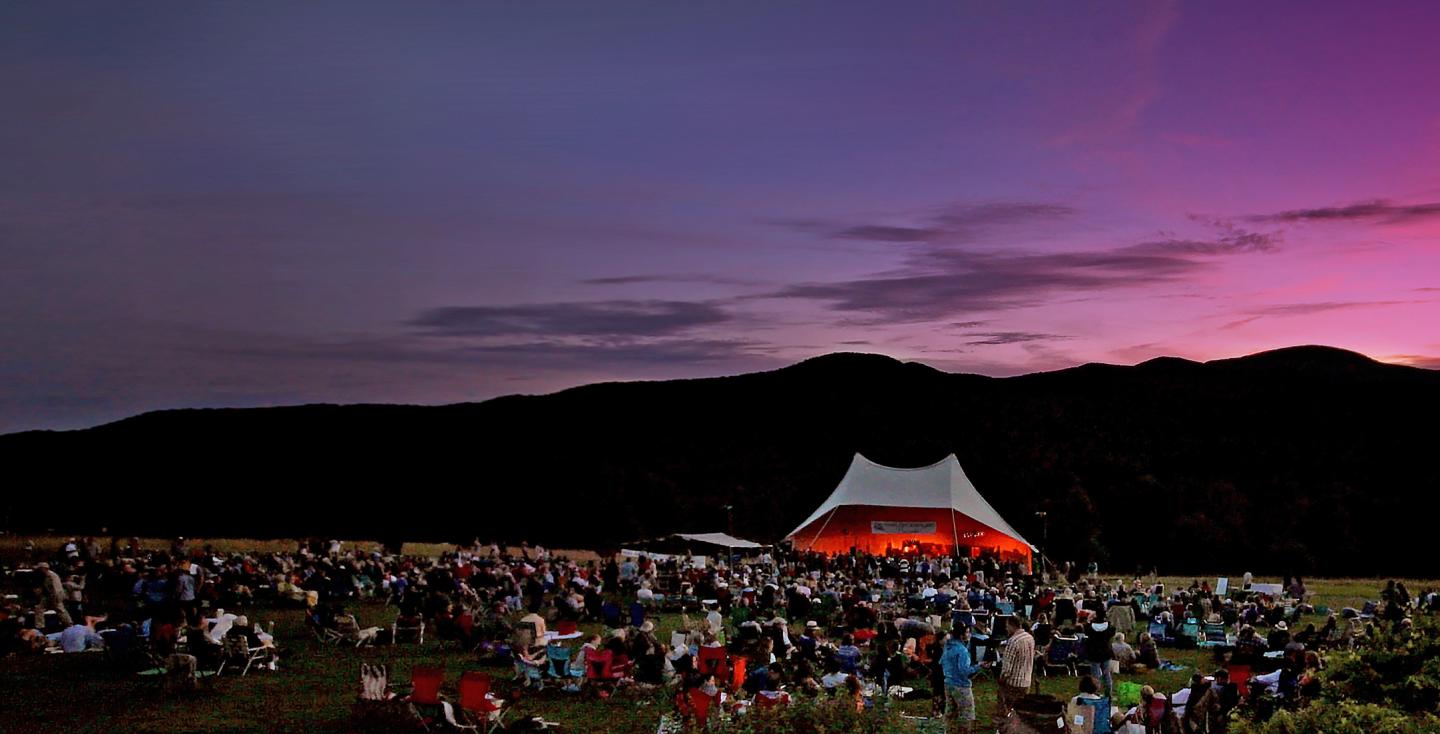 Outdoor concert at dusk with a crowd and a lit tent stage against a mountain backdrop.