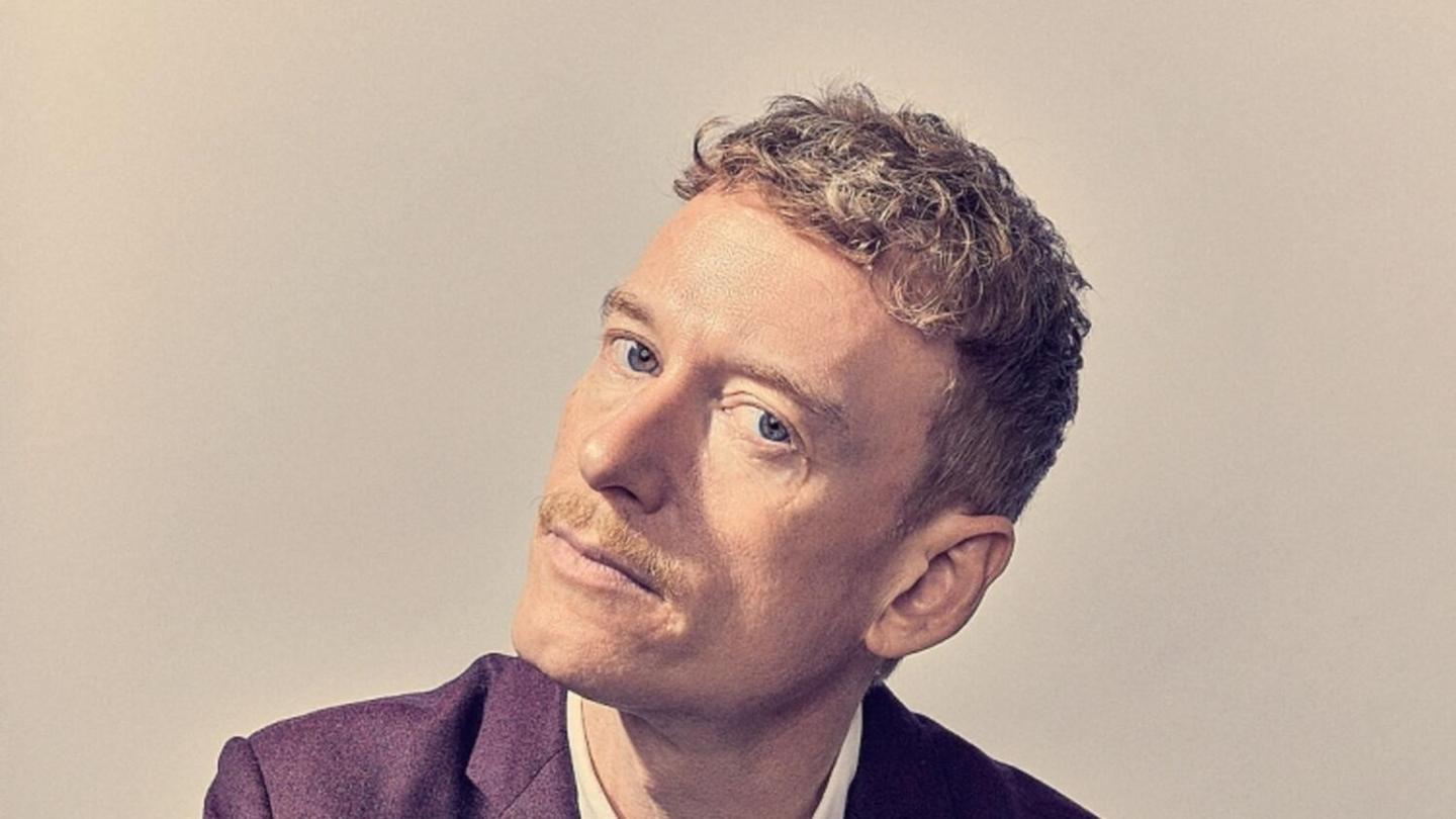 A person with curly hair and a mustache wearing a suit, tilting their head slightly.
