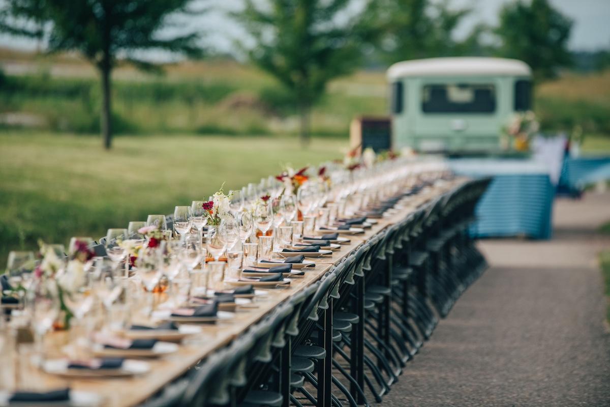 Outdoor dining setup with long tables, wine glasses, and a vintage van.