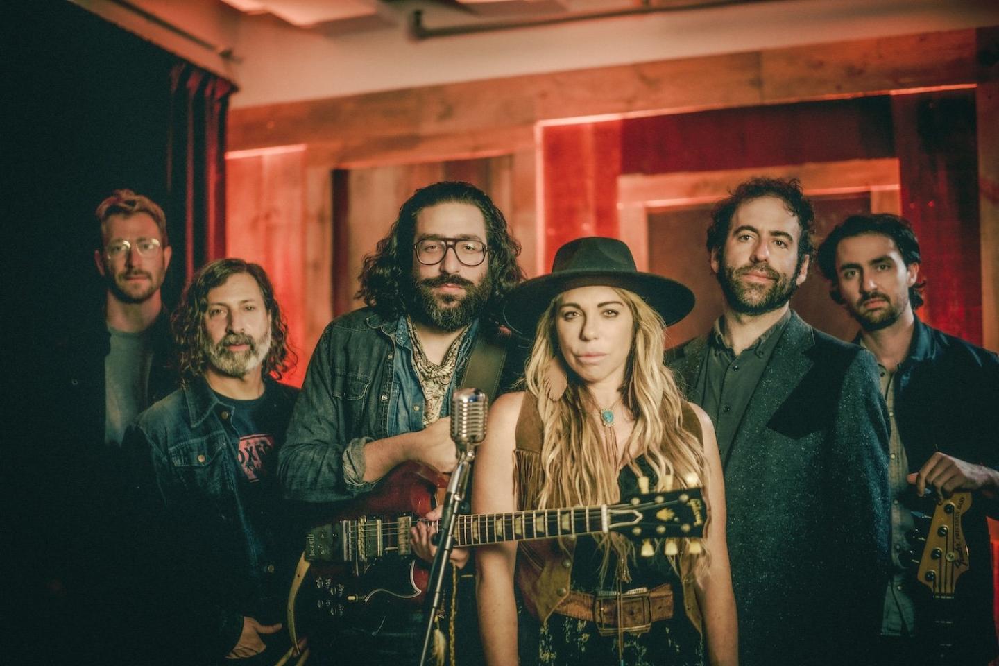 Band posing with guitars in a warmly lit studio, featuring six members.