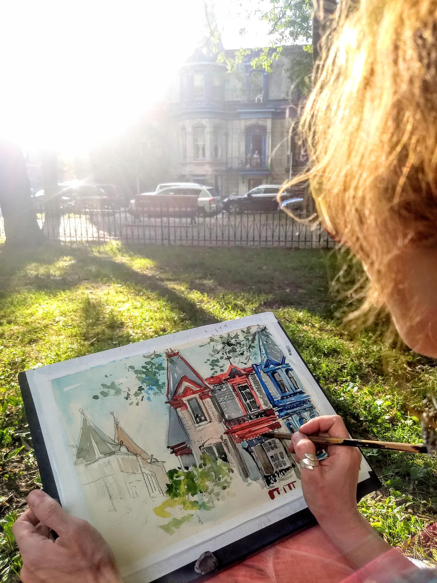 Artist painting vibrant houses in sunlight, seated in a grassy yard.