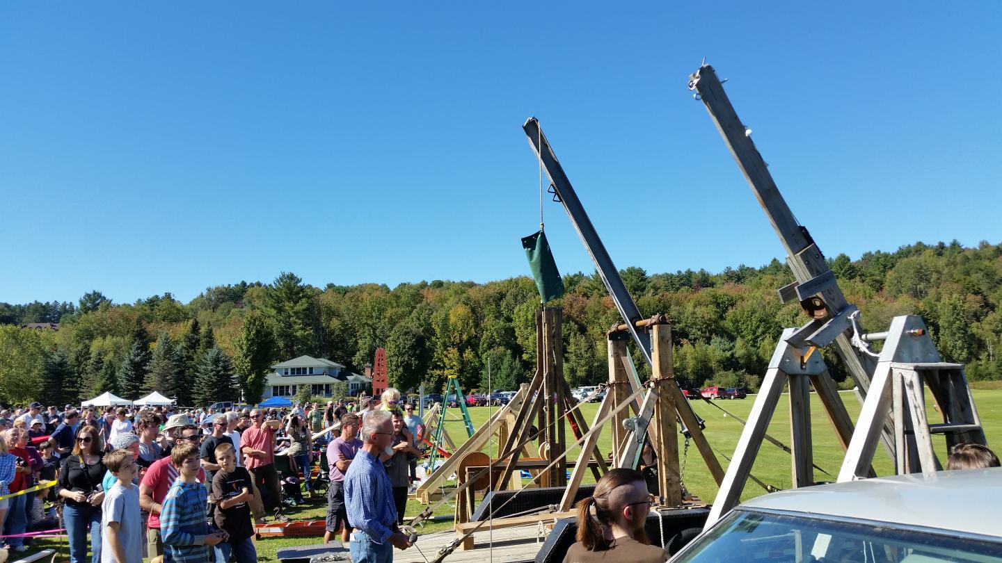 Trebuchets on a field with a crowd watching under a clear blue sky.