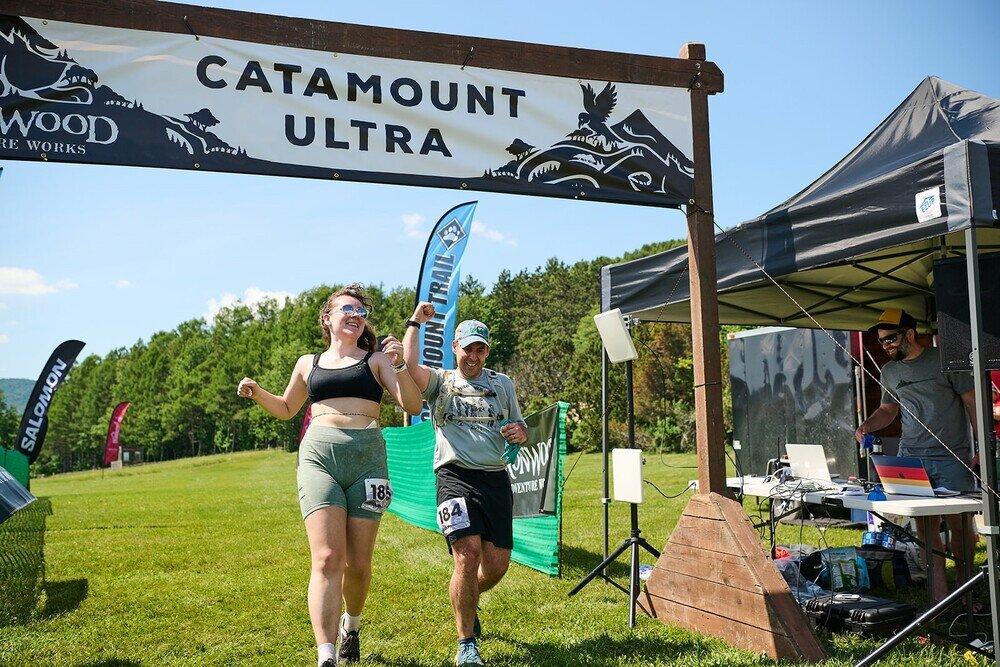 Runners cross finish line under Catamount Ultra banner, smiling, in outdoor setting.