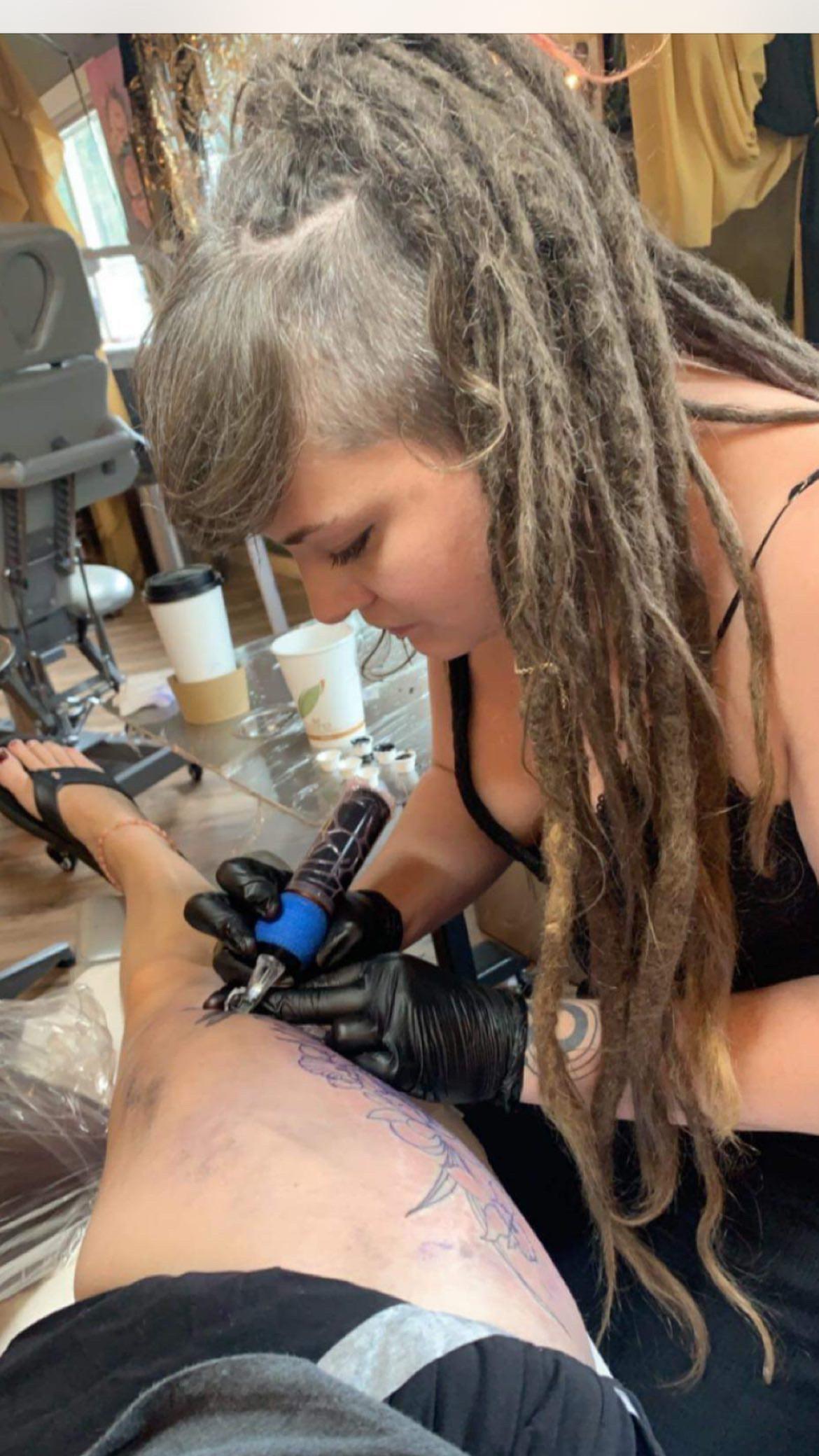 Tattoo artist with long dreadlocks tattooing a person's leg.