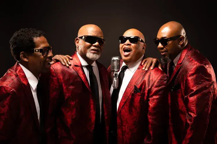 Four men in red suits singing, wearing sunglasses, with a microphone on a dark background.