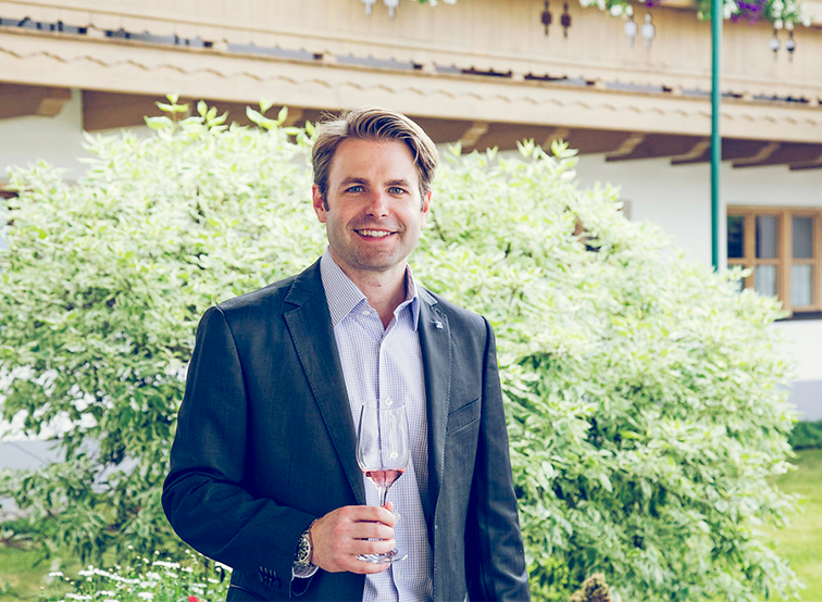 Man in blazer smiling, holding wineglass, lush greenery background.