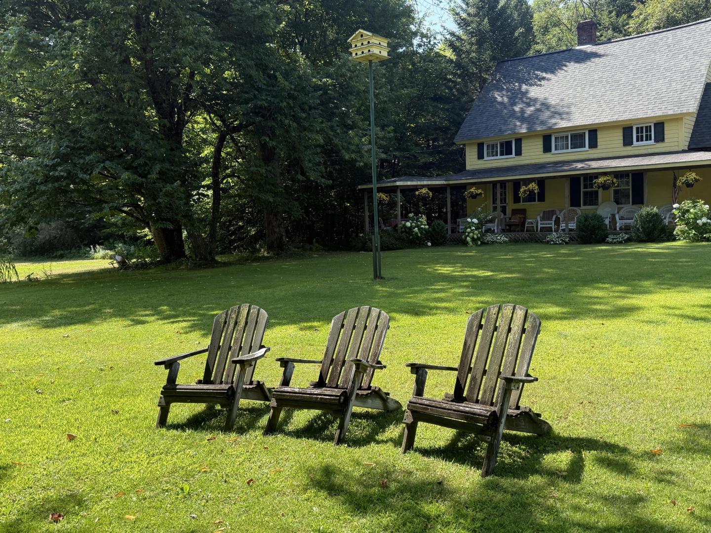 Three wooden chairs on lawn in front of a yellow house with a porch.