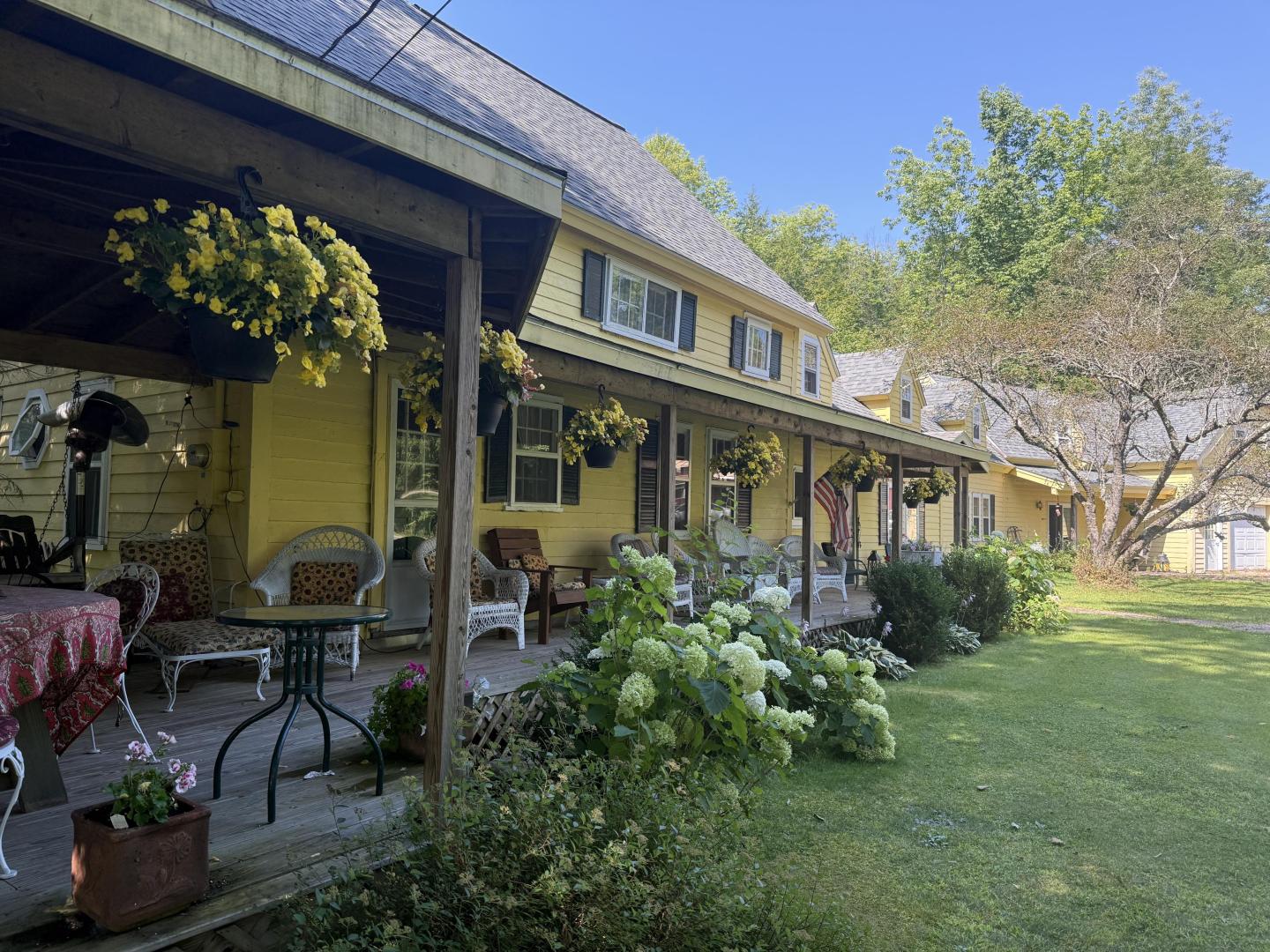 Charming yellow house with a long porch, flowers, and greenery on a sunny day.