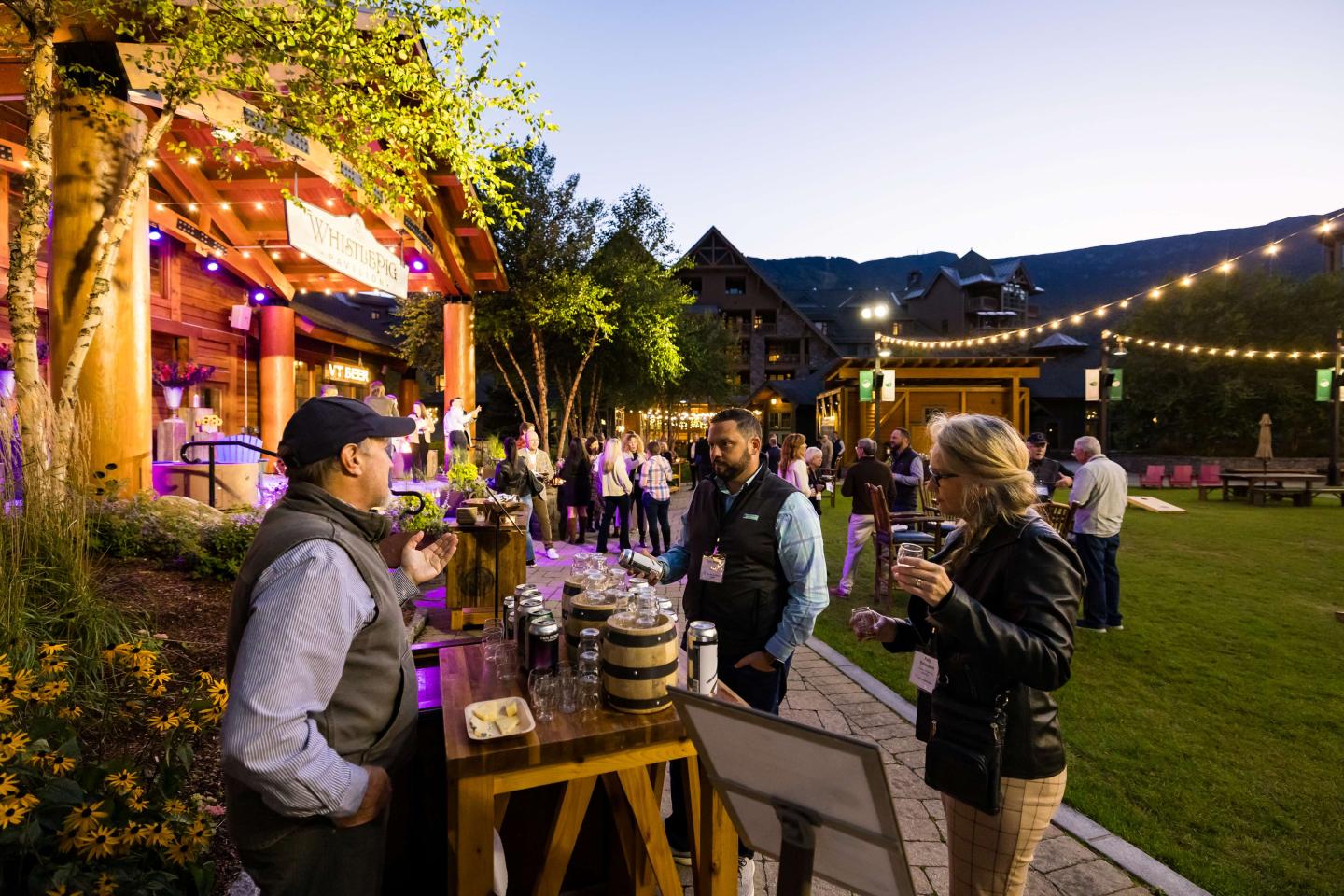 Outdoor gathering at dusk with people socializing near a wooden bar.