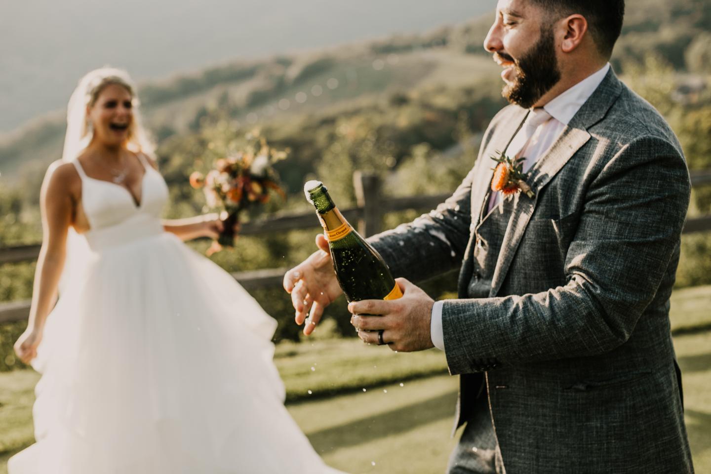 Man opening champagne with bride laughing, outdoors on a sunny day.