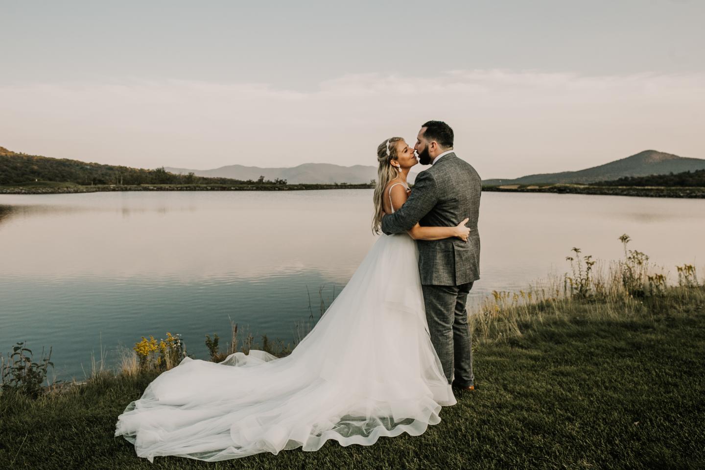 Bride and groom embrace by a serene lake, mountains in the background.