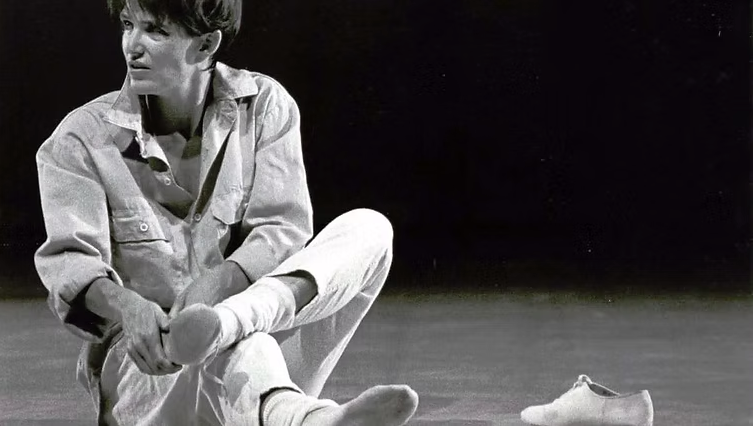 Person sitting, adjusting shoe on a stage with a contemplative expression.