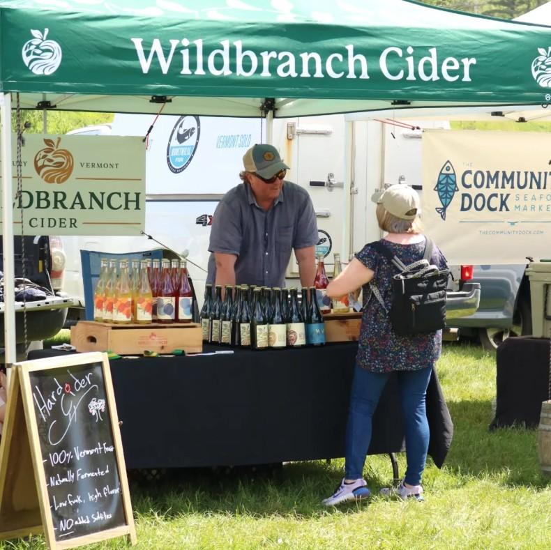 Vendor stall with cider bottles under a green tent, two people talking.
