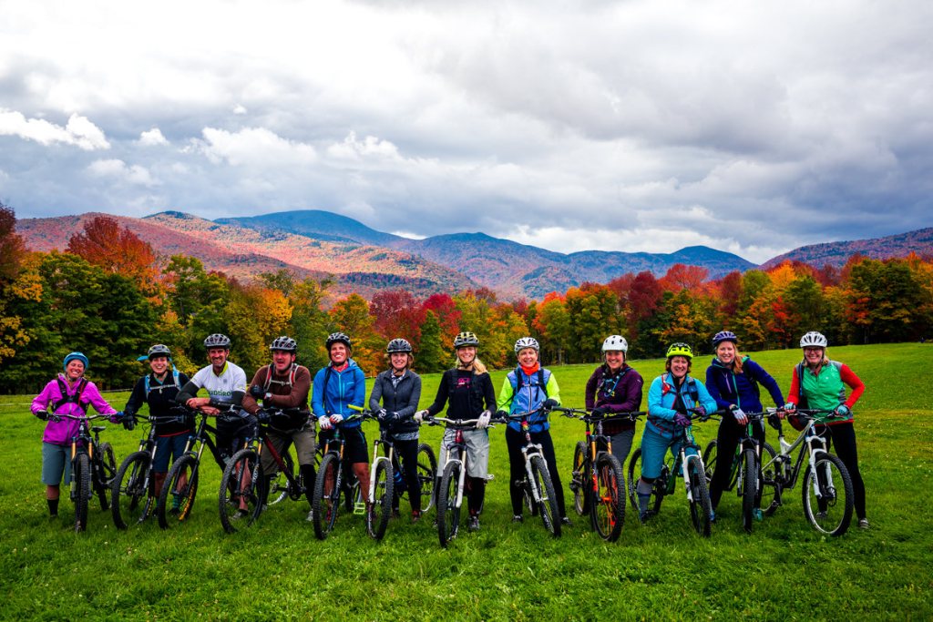 A group of cyclists stand with bikes in a field, autumn trees and mountains behind.