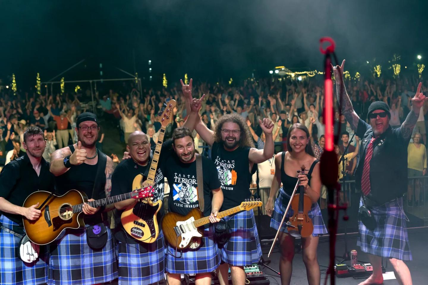 Band on stage in kilts, smiling with hands raised, large crowd in background.