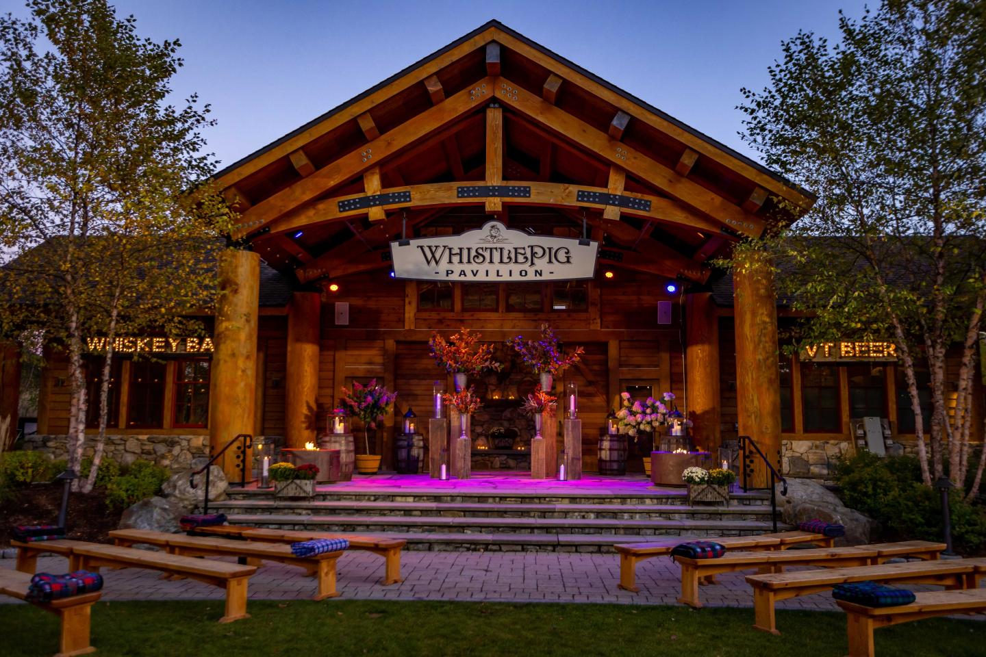 Wooden pavilion at dusk with stage and floral decorations, set in a garden.