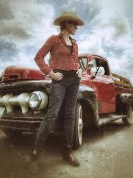 Cowboy in hat stands by a red vintage truck under a cloudy sky.