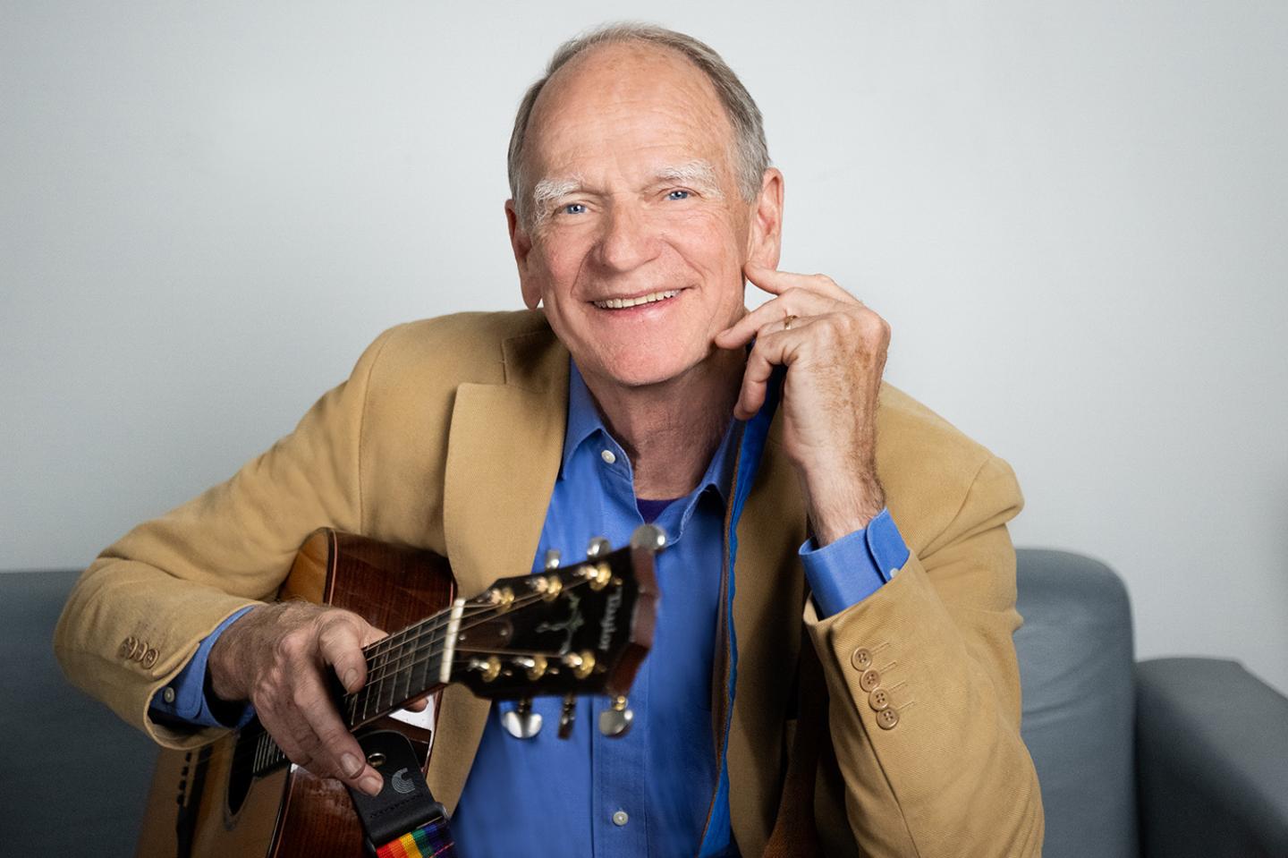 Smiling man with a guitar, sitting on a couch, wearing a blue shirt and tan blazer.