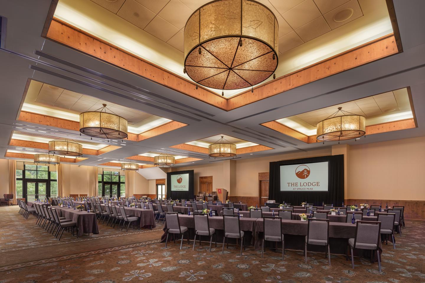 Conference room with rows of chairs facing two large screens, warm lighting overhead.