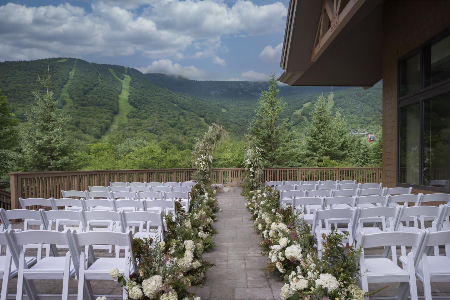 Outdoor wedding aisle with white chairs and floral arrangements, facing green mountains.