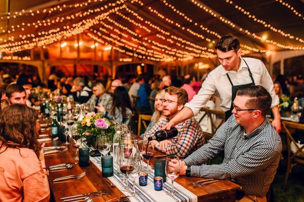 Server pouring wine for guests at a festive, warmly lit dinner party.