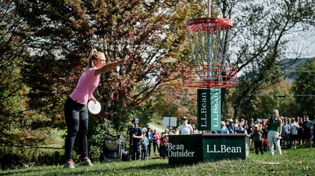 Disc golfer in pink shirt putting towards basket, with crowd and trees in background.