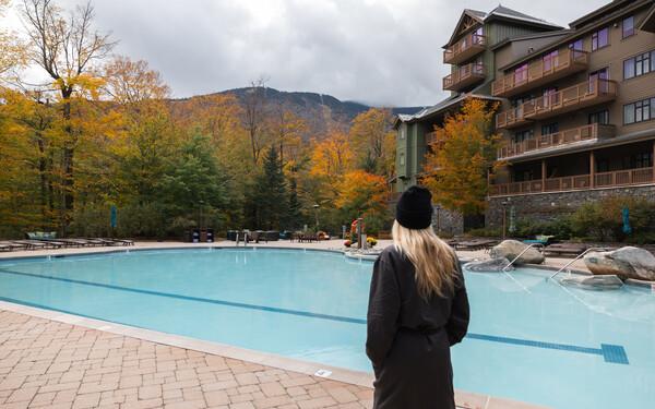 Person in a coat and hat overlooking a pool, surrounded by autumn trees and a building.