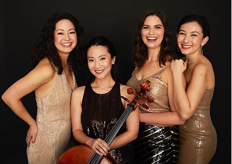 Four women in elegant dresses pose with a cello, smiling against a dark background.