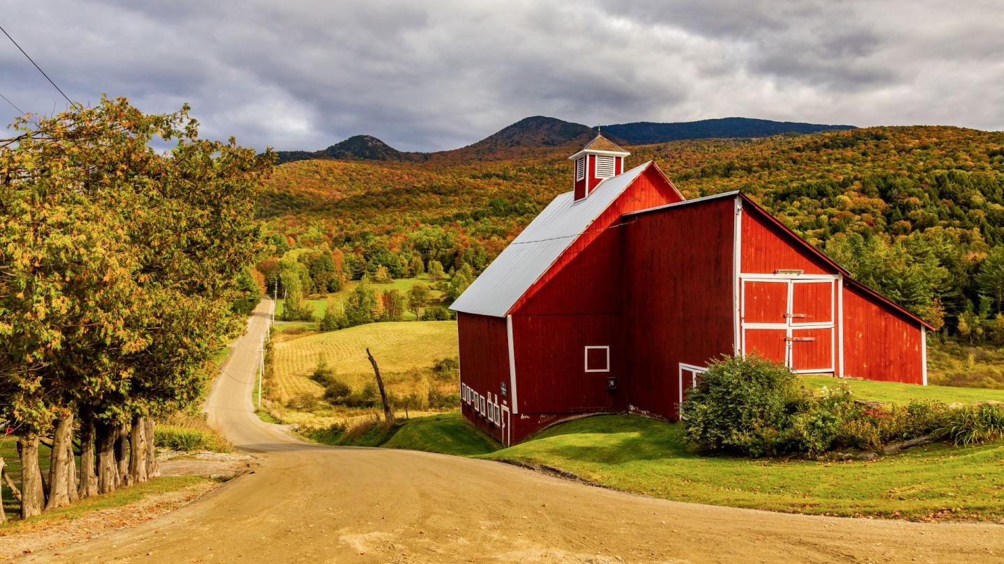 Red barn on a dirt road with autumn trees and mountains in the background.