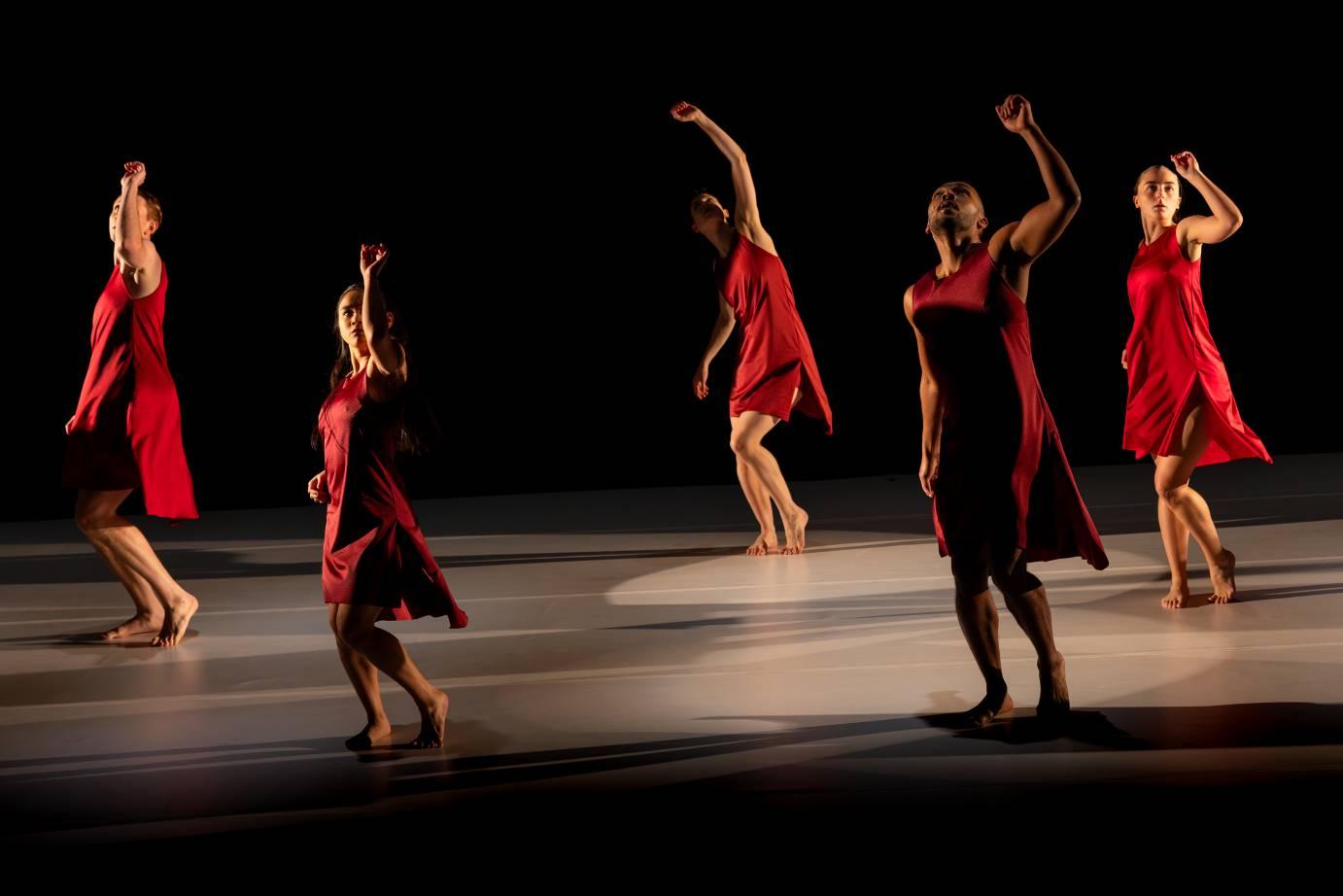Five dancers in red dresses perform on a dimly lit stage.
