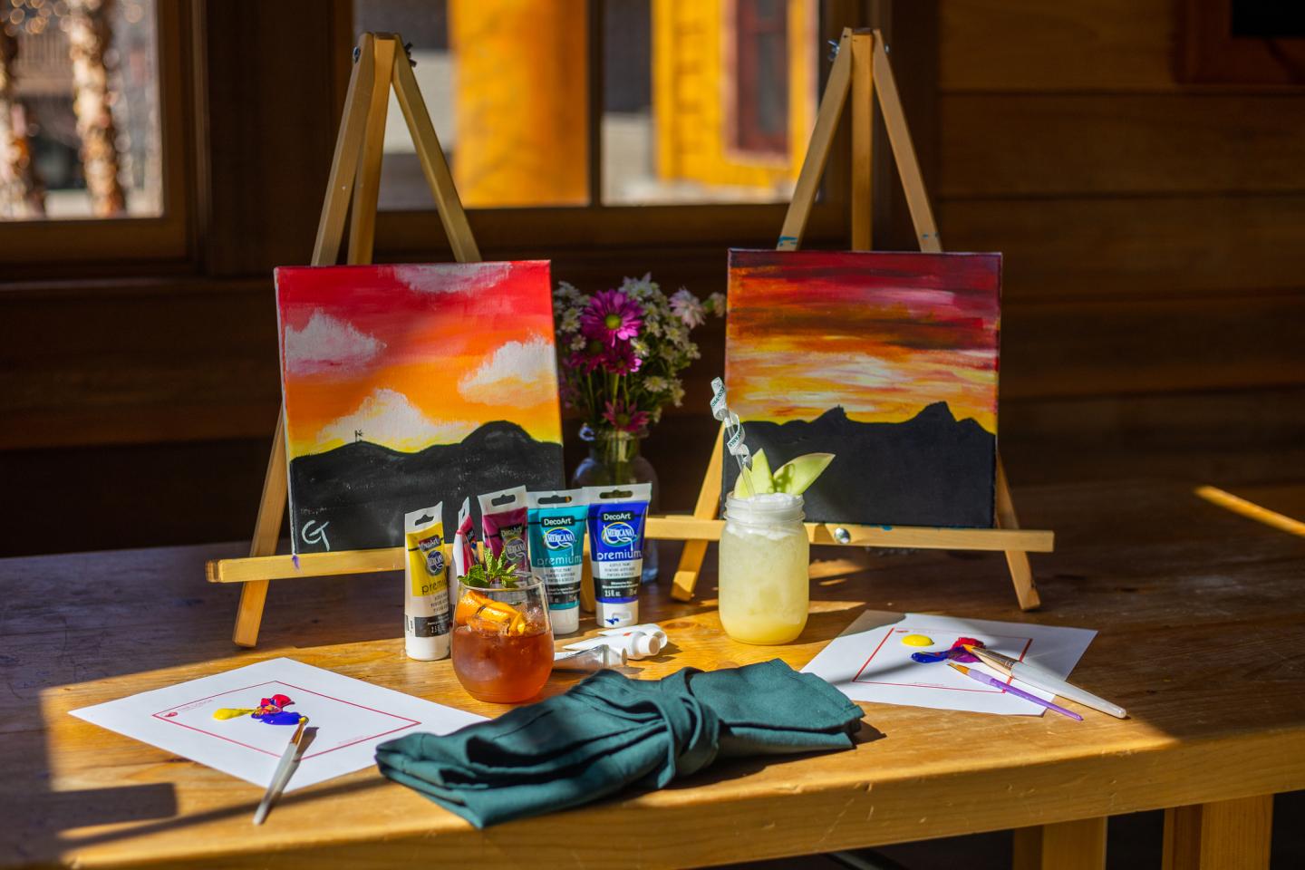 Sunset paintings on easels, art supplies on a wooden table.