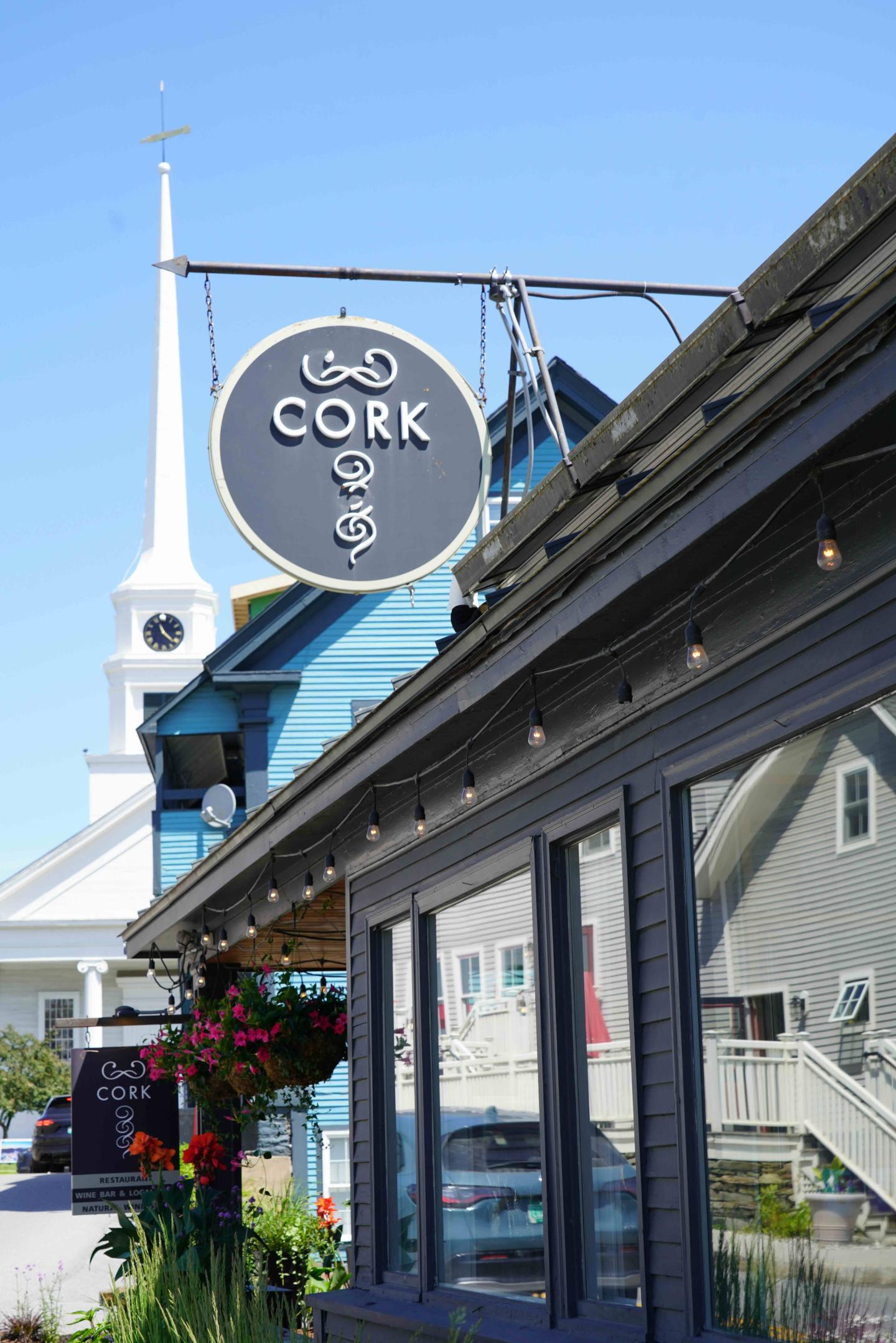 Sign for Cork Restaurant, near a white church steeple, under a clear blue sky.